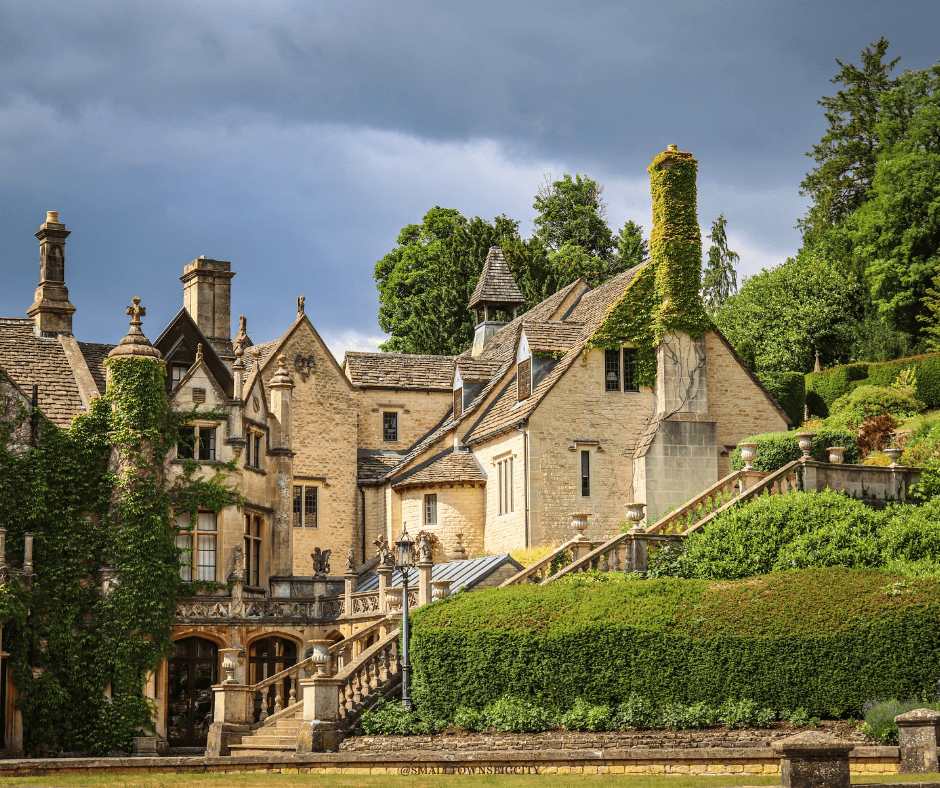 A grand honey-stone Cotswolds manor house with gabled roofs, ivy-clad chimneys, stone balustrade staircases, and manicured hedges under a moody grey sky.